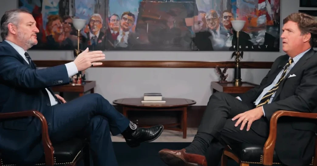Ted Cruz (left), in a suit, gestures while speaking to Tucker Carlson (right), also in a suit, who listens intently. They are seated facing each other during an interview, with framed portraits in the background.