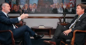Ted Cruz (left), in a suit, gestures while speaking to Tucker Carlson (right), also in a suit, who listens intently. They are seated facing each other during an interview, with framed portraits in the background.
