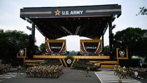 U.S. Army soldiers marching in formation during a large-scale parade in front of a massive stage branded with the U.S. Army logo. M1 Abrams tanks are on display, and large screens show the logo of the sponsor, Oracle.
