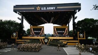 U.S. Army soldiers marching in formation during a large-scale parade in front of a massive stage branded with the U.S. Army logo. M1 Abrams tanks are on display, and large screens show the logo of the sponsor, Oracle.