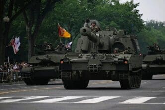 An American M109A7 Paladin self-propelled howitzer, flanked by two M1 Abrams tanks, drives down a street during a military parade as soldiers look out from the hatches and spectators watch from the sidewalk.