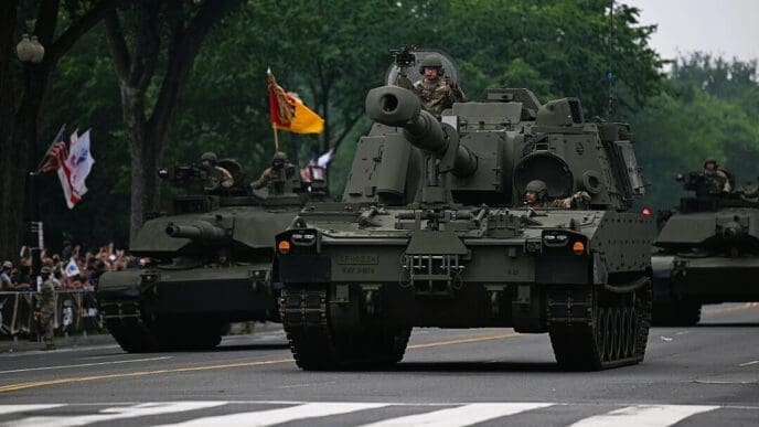 An American M109A7 Paladin self-propelled howitzer, flanked by two M1 Abrams tanks, drives down a street during a military parade as soldiers look out from the hatches and spectators watch from the sidewalk.