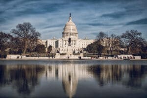 The U.S. Capitol Building, with its iconic dome and neoclassical architecture, is perfectly reflected in the Capitol Reflecting Pool in Washington, D.C., framed by bare trees under a dramatic sky.