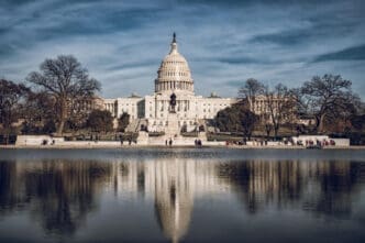 The U.S. Capitol Building, with its iconic dome and neoclassical architecture, is perfectly reflected in the Capitol Reflecting Pool in Washington, D.C., framed by bare trees under a dramatic sky.