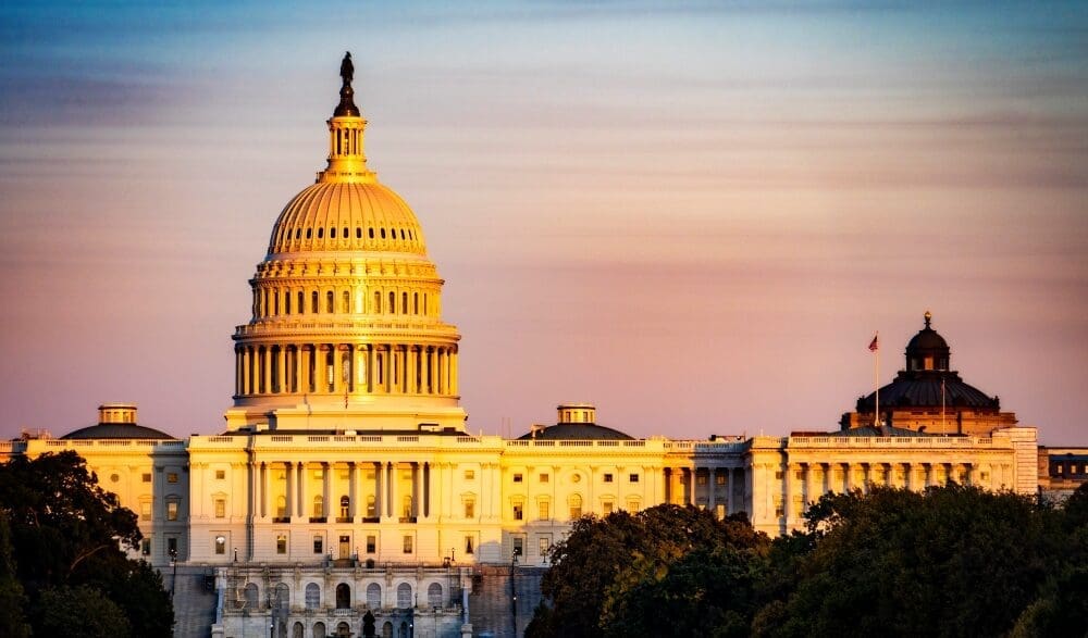 The U.S. Capitol Building is bathed in warm, golden light during sunset or sunrise, with its iconic dome, columns, and expansive wings visible against a pastel-colored sky.