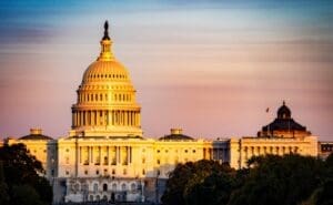 The U.S. Capitol Building is bathed in warm, golden light during sunset or sunrise, with its iconic dome, columns, and expansive wings visible against a pastel-colored sky.