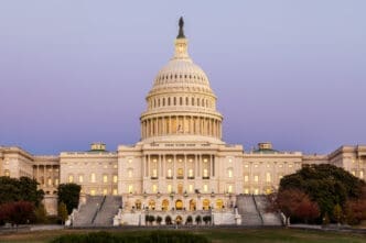 The U.S. Capitol Building in Washington, D.C., is brightly illuminated at dusk, showcasing its iconic dome, grand steps, and classical architecture against a purple and blue twilight sky.