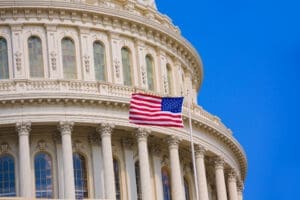 A close-up view of the U.S. Capitol Building's white dome, with an American flag flying prominently on a flagpole in front of its classical architecture, under a clear blue sky.