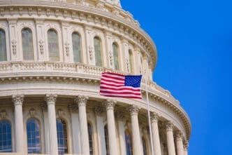 A close-up view of the U.S. Capitol Building's white dome, with an American flag flying prominently on a flagpole in front of its classical architecture, under a clear blue sky.