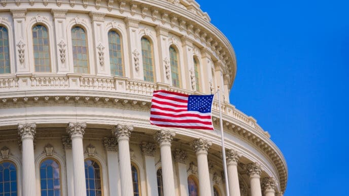A close-up view of the U.S. Capitol Building's white dome, with an American flag flying prominently on a flagpole in front of its classical architecture, under a clear blue sky.