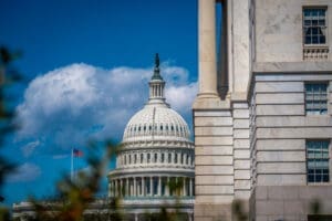 A close-up view of the U.S. Capitol Building's white dome and a portion of its neoclassical facade, with the American flag flying nearby, framed by another stone building and tree branches under a blue sky with white clouds.