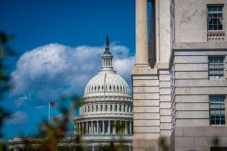 A close-up view of the U.S. Capitol Building's white dome and a portion of its neoclassical facade, with the American flag flying nearby, framed by another stone building and tree branches under a blue sky with white clouds.