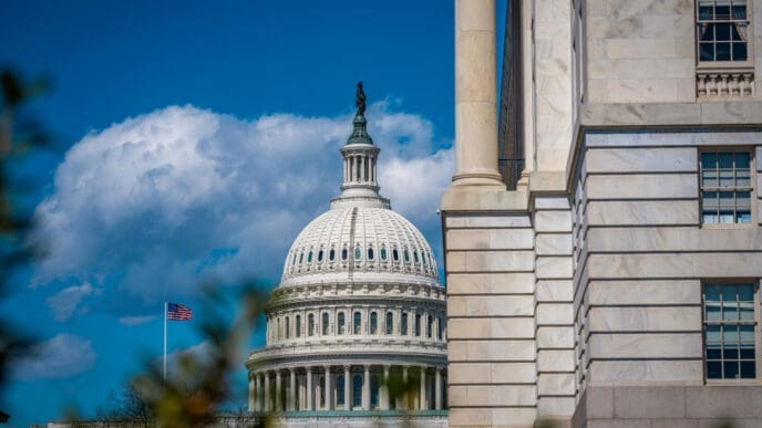 A close-up view of the U.S. Capitol Building's white dome and a portion of its neoclassical facade, with the American flag flying nearby, framed by another stone building and tree branches under a blue sky with white clouds.