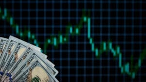 A fan of one-hundred-dollar bills is shown in the foreground against a blurry, glowing green financial stock chart in the background that shows a sharp downward trend.