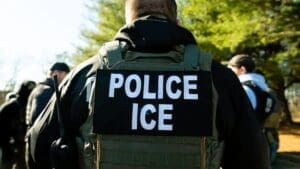 A view from behind of a U.S. Immigration and Customs Enforcement (ICE) officer wearing a tactical vest with "POLICE" and "ICE" written in large white letters on the back during a field operation.