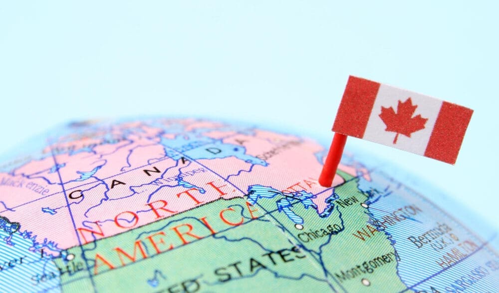 A close-up photograph of a globe showing the map of North America, with the border between Canada and the United States prominently visible. A Canadian flag is pinned into the map near this border, symbolizing cross-border trade.
