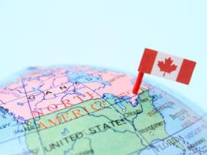 A close-up photograph of a globe showing the map of North America, with the border between Canada and the United States prominently visible. A Canadian flag is pinned into the map near this border, symbolizing cross-border trade.