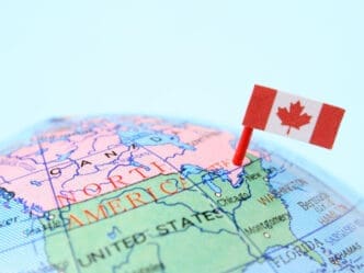 A close-up photograph of a globe showing the map of North America, with the border between Canada and the United States prominently visible. A Canadian flag is pinned into the map near this border, symbolizing cross-border trade.