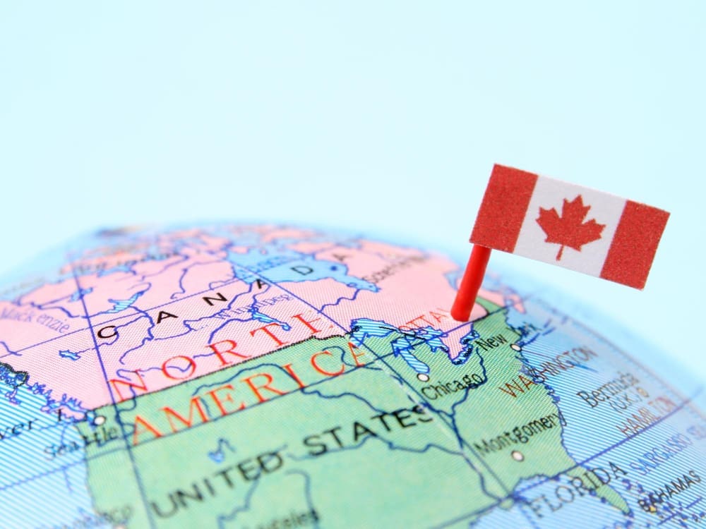 A close-up photograph of a globe showing the map of North America, with the border between Canada and the United States prominently visible. A Canadian flag is pinned into the map near this border, symbolizing cross-border trade.