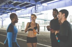 A diverse group of four friends in running gear stand together talking and smiling under a city bridge after a workout, holding sports drinks to rehydrate.