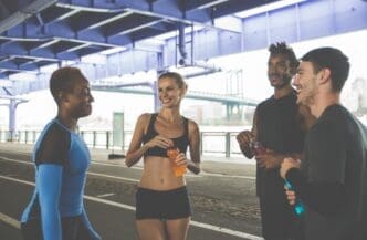 A diverse group of four friends in running gear stand together talking and smiling under a city bridge after a workout, holding sports drinks to rehydrate.