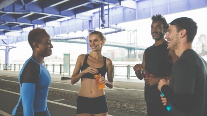 A diverse group of four friends in running gear stand together talking and smiling under a city bridge after a workout, holding sports drinks to rehydrate.