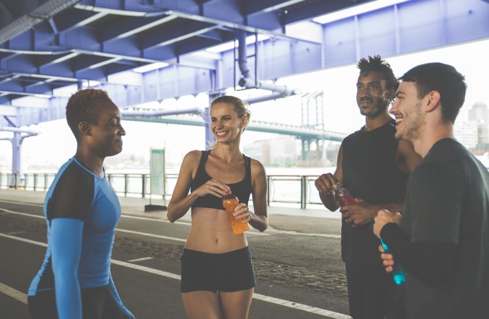 A diverse group of four friends in running gear stand together talking and smiling under a city bridge after a workout, holding sports drinks to rehydrate.