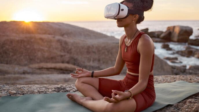 A woman, who appears to be Black, sits cross-legged in a yoga pose on a mat on rocky terrain by the sea, wearing a white virtual reality (VR) headset, with a bright sunset in the background.