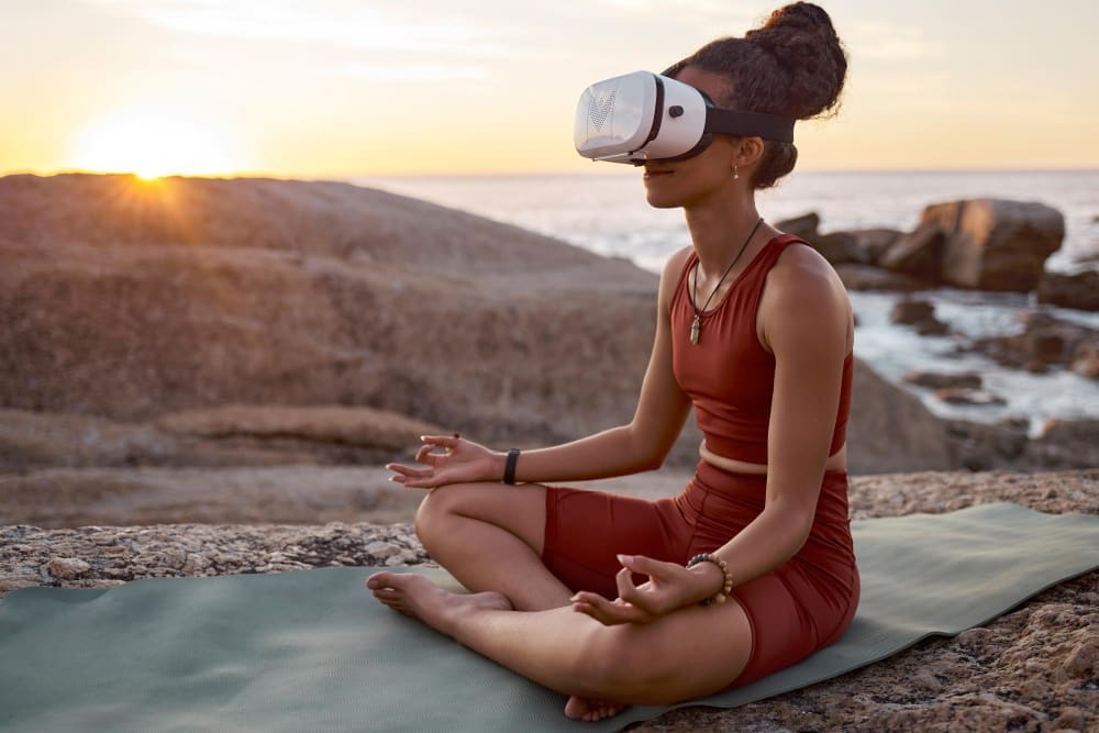 A woman, who appears to be Black, sits cross-legged in a yoga pose on a mat on rocky terrain by the sea, wearing a white virtual reality (VR) headset, with a bright sunset in the background.