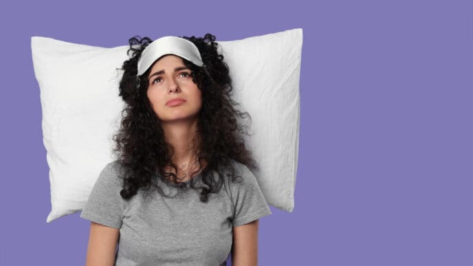 A young woman with curly hair and a sleep mask on her forehead is lying on a white pillow, looking upwards with a tired and unhappy expression against a solid purple background.