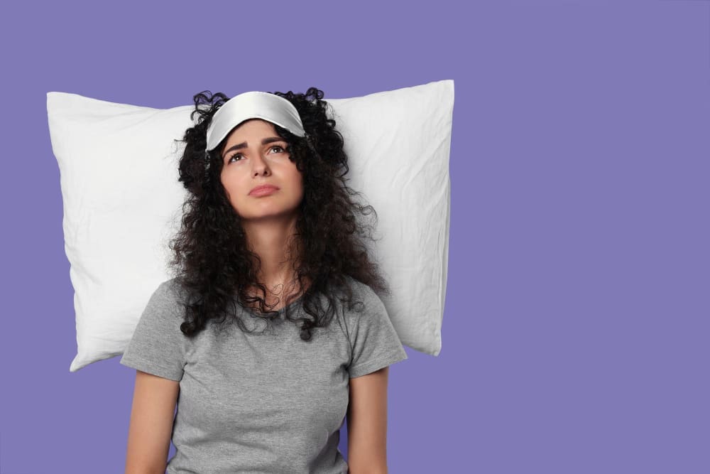 A young woman with curly hair and a sleep mask on her forehead is lying on a white pillow, looking upwards with a tired and unhappy expression against a solid purple background.