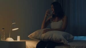 A young woman, who appears to be of East Asian descent, sits up in bed in a dark room, drinking a glass of water. A lamp is lit on her bedside table next to a bottle of water and a pill bottle.