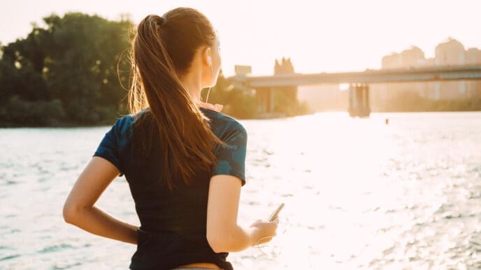 A woman in athletic clothes, seen from behind with her hair in a ponytail, looking out over a body of water towards a city skyline and a bridge during a bright sunrise.