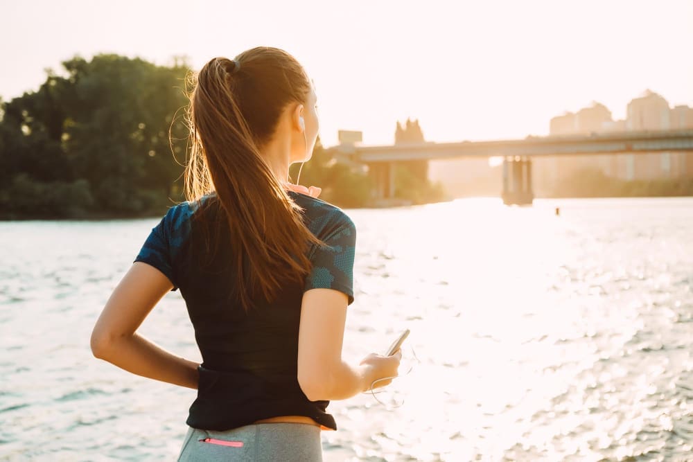 A woman in athletic clothes, seen from behind with her hair in a ponytail, looking out over a body of water towards a city skyline and a bridge during a bright sunrise.