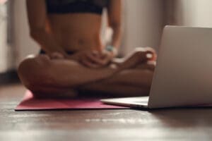 A person in athletic wear sits in a cross-legged yoga or meditation pose on a pink mat, with a silver laptop open on the wooden floor in front of them.