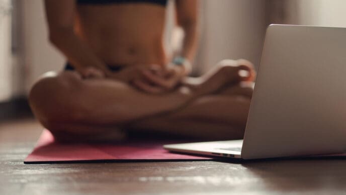 A person in athletic wear sits in a cross-legged yoga or meditation pose on a pink mat, with a silver laptop open on the wooden floor in front of them.