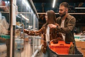 A side view of a young couple, who appear to be White, shopping in a supermarket. The woman is opening the glass door to a refrigerated aisle while the man stands behind her and points at a product.
