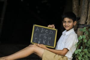 A smiling young boy, who appears to be of Indian descent and is wearing a school uniform, sits on the ground while holding up a small chalkboard with the letters of the alphabet written on it.