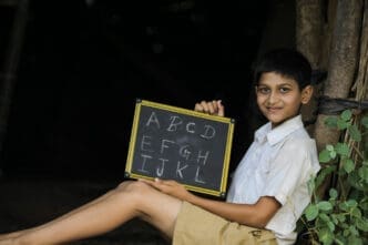 A smiling young boy, who appears to be of Indian descent and is wearing a school uniform, sits on the ground while holding up a small chalkboard with the letters of the alphabet written on it.