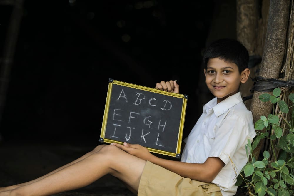 A smiling young boy, who appears to be of Indian descent and is wearing a school uniform, sits on the ground while holding up a small chalkboard with the letters of the alphabet written on it.
