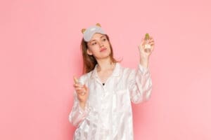 A young woman wearing pajamas and a sleep mask on her forehead holds a spray bottle while posing against a solid pink background.