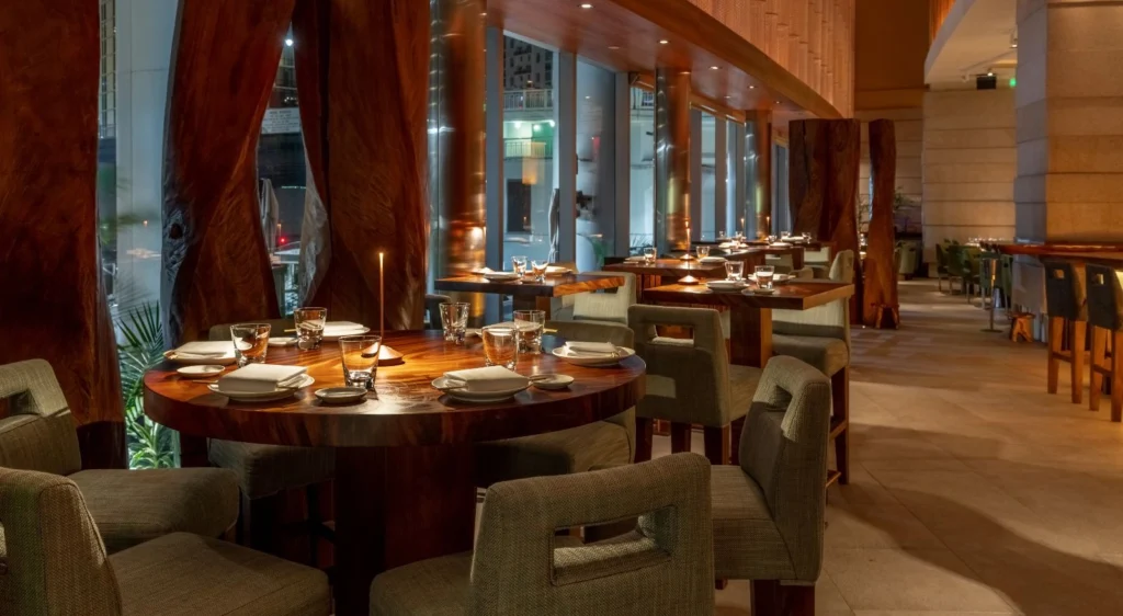 The elegant interior of Zuma Miami restaurant at night, showing multiple round wooden dining tables set for guests, with distinctive dark wood panels, modern seating, and large windows looking out onto a city view.