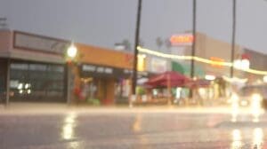 A blurry, low-angle photo of a city street at night or dusk during heavy rain, showing bright reflections of streetlights and car headlights on the wet road, with blurred storefronts and palm trees in the background.