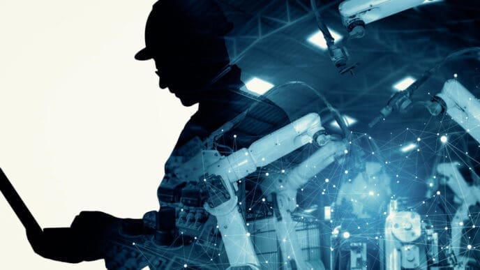 A silhouette of an engineer in a hard hat working on a laptop, overlaid with a blue-toned digital network and robotic arms, symbolizing AI's disruption of industrial sectors.