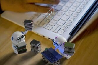 Two small, white humanoid robots with glasses are diligently sorting through stacks of miniature books and a tiny laptop on a wooden surface, with human hands typing on a larger laptop in the background and a glowing chat icon floating above.