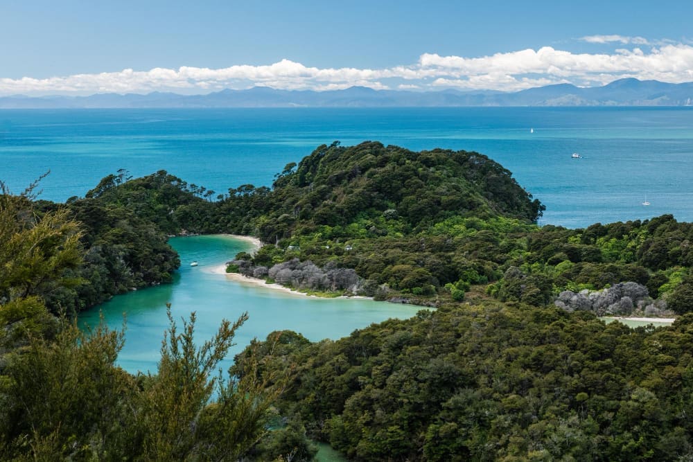 An aerial view of Abel Tasman National Park, showing lush green forested peninsulas enclosing a calm, turquoise bay with a small boat, extending into the vast blue ocean with distant mountains under a partly cloudy sky.