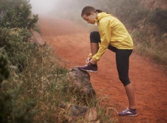 A woman in a yellow jacket and leggings tying her trail running shoe on a log on a dirt path, in a misty outdoor setting.