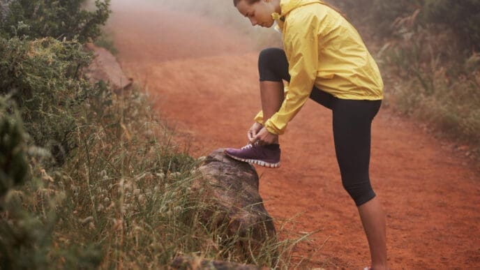 A woman in a yellow jacket and leggings tying her trail running shoe on a log on a dirt path, in a misty outdoor setting.