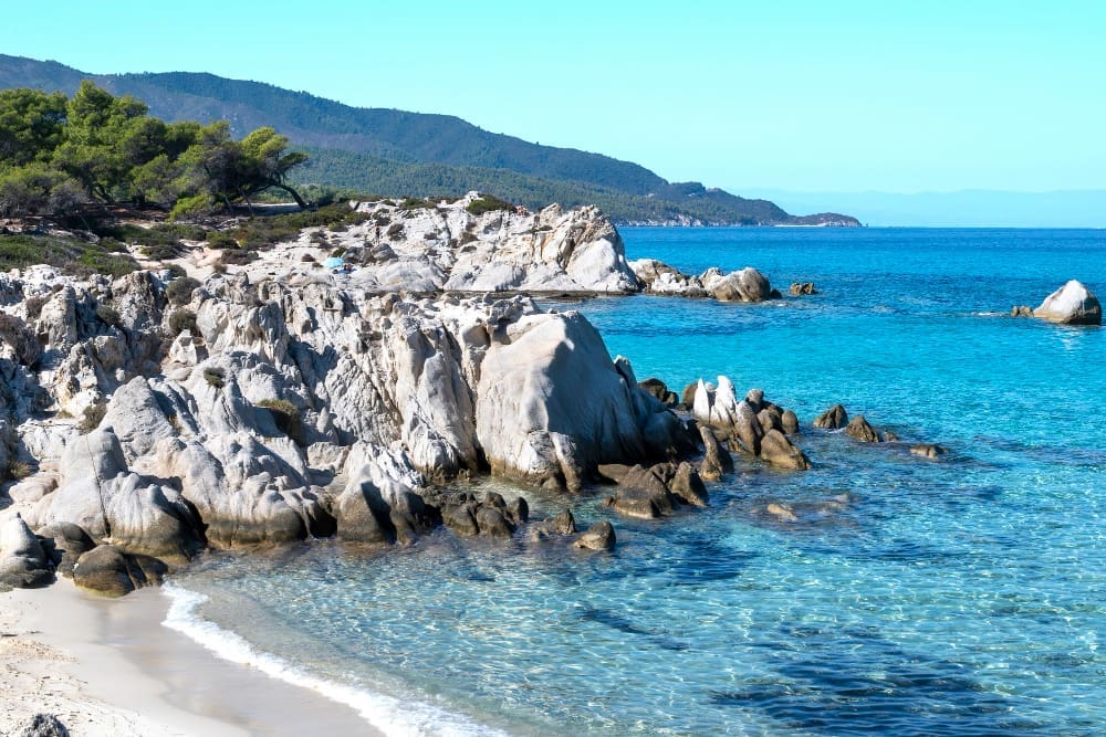A pristine Aegean Sea coast in Greece, with crystal-clear turquoise water lapping a white sand beach. Dramatic light-colored rock formations, green trees, and distant mountains create a scenic landscape.