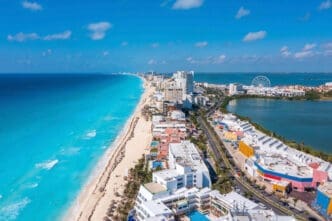 An expansive aerial view of Cancun, Mexico, showing a long stretch of white sand beach and turquoise ocean on the left, luxury hotels and resorts lining a narrow land strip, and a calm lagoon with a Ferris wheel on the right, all under a clear blue sky.